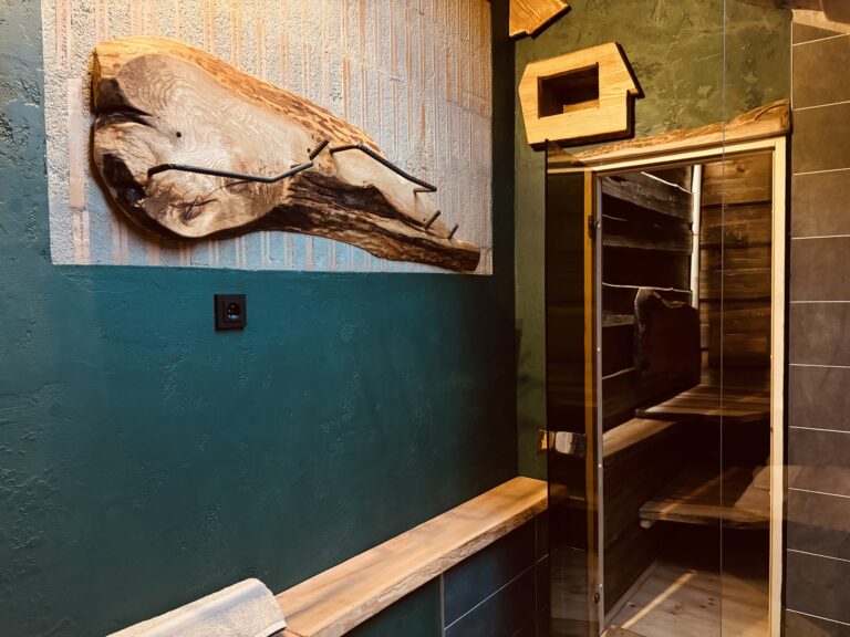 Detail vstupu do sauny s prosklenými dveřmi, tmavě zelenou stěnou a unikátním dřevěným věšákem z masivu.